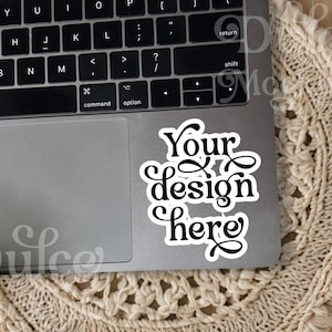 May include: A silver laptop computer with a white sticker on the trackpad that says "Your design here". The laptop is sitting on a beige woven mat.