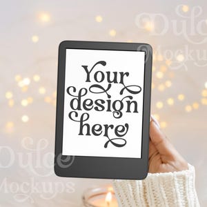 May include: A black e-reader with a white screen displaying the text "Your design here" in a decorative font. The e-reader is held in a hand wearing a white knitted sweater against a blurred background of white and gold lights.