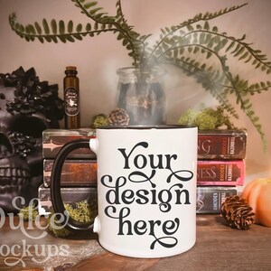 May include: White ceramic mug with a black rim and handle. The mug has the text "Your design here" in black cursive lettering. The mug is sitting on a wooden table with a skull, books, a candle, and a fern in the background.