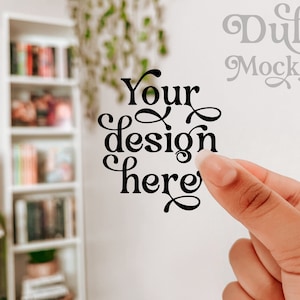May include: A hand holds up a white sticker with black text that says "Your design here". The text is in a decorative font. The background is blurred and shows a bookshelf with books.