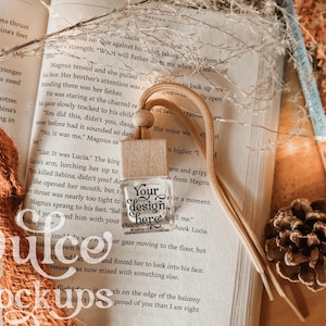 May include: A small, square, clear glass bottle with a light-colored wooden cap and a tan cord for hanging. The front of the bottle features the black printed text "Your design here". The bottle is positioned on an open book page.