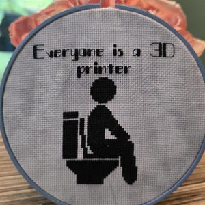 May include: A circular embroidery hoop containing a light gray fabric with black cross-stitched text that reads Everyone is a 3D printer, accompanied by a black cross-stitched icon of a stick figure sitting on a toilet, framed in a blue plastic hoop.