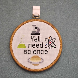 以下が含まれることがあります： 茶色の木製の枠が付いた白い刺繍枠。刺繍はビーカー、顕微鏡、原子、惑星、そして「y'all need science」という文字を表しています。