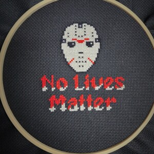 Può includere: Un cerchio da ricamo a punto croce con un'immagine in bianco e nero di una maschera da hockey e il testo "No Lives Matter" in rosso e bianco.