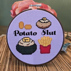 Op de afbeelding: Een borduurring met kruissteek met een lichtpaarse achtergrond. Het ontwerp toont verschillende afbeeldingen met aardappelthema, waaronder friet en een gepofte aardappel, met de woorden "Potato Slut" in zwart.