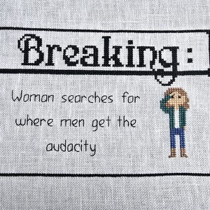 May include: A cross-stitch design with a black border and white background. The text "Breaking:" is at the top of the design. The text "Woman searches for where men get the audacity" is below the title. A cartoon image of a woman with brown hair is on the right side of the design.