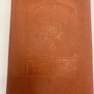 Pode incluir: Um livro vintage, castanho-avermelhado, intitulado "French and English First Book". A capa apresenta elementos decorativos em relevo, incluindo um emblema central e bordas ornamentadas. A capa do livro é de uma cor quente e sólida.