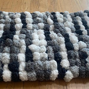 May include: A rectangular pom-pom blanket in white, grey, and black. The handmade blanket is made of soft, fluffy yarn, creating a textured surface. The pom-poms are evenly spaced, giving the blanket a cosy appearance.