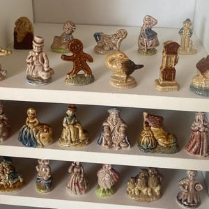 Puede incluir: Una colección de figuras de cerámica vintage expuestas en estantes blancos. Las figuras representan varios personajes y objetos, incluyendo un gato tocando el violín, un hombre de jengibre y una casa. Las figuras son de varios colores y tamaños.