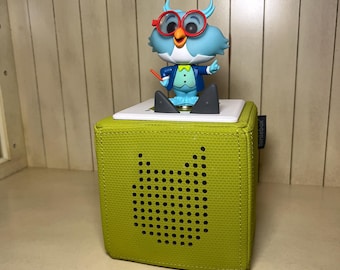 May include: A lime green fabric-covered speaker with a cat-shaped speaker grill. A blue owl figurine with red spectacles and a conductor's baton sits on top. The speaker has the word "toniebox" printed on the side.