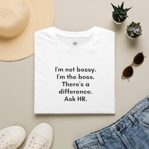 May include: A white crew neck t-shirt featuring the black printed text: I'm not bossy. I'm the boss. There's a difference. Ask HR. The shirt is displayed alongside a straw hat, two small potted succulents, brown sunglasses, and white sneakers.
