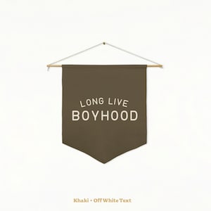 May include: A dark olive green fabric wall banner featuring the off-white text LONG LIVE BOYHOOD printed in a sans-serif font. The banner is suspended from a horizontal wooden dowel with a thin white string attached to the ends for hanging.