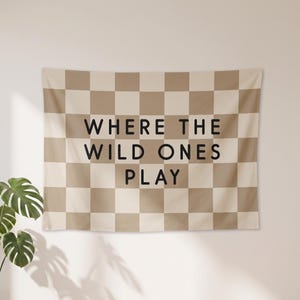 May include: A rectangular wall tapestry featuring a beige and cream chequered pattern. The text "WHERE THE WILD ONES PLAY" is printed in bold, black, sans-serif capital letters across the centre of the fabric, centred horizontally.