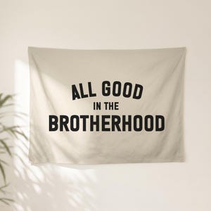 May include: A rectangular beige wall tapestry featuring the black text ALL GOOD IN THE BROTHERHOOD printed in a bold, sans-serif font. The top line is arched, while the middle and bottom lines are straight, centred on the fabric surface.