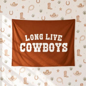 May include: A rectangular wall tapestry in a solid rust-orange colour featuring the white text LONG LIVE COWBOYS in a bold, serif font. The background pattern consists of small, repeating illustrations of cowboy hats, boots, lassos, and stars.