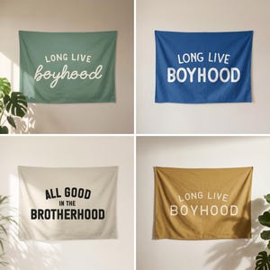 May include: Four rectangular fabric wall tapestries. One is sage green with white text reading LONG LIVE boyhood. One is blue with white text reading LONG LIVE BOYHOOD. One is cream with black text reading ALL GOOD IN THE BROTHERHOOD. One is mustard yellow with white text reading LONG LIVE BOYHOOD.