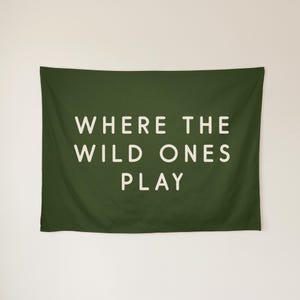 May include: A rectangular dark olive green wall tapestry featuring the text WHERE THE WILD ONES PLAY in a clean, off-white sans-serif font centred on the fabric. The design is printed clearly against the solid dark green background.