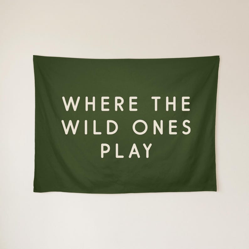 May include: A rectangular dark olive green wall tapestry featuring the text WHERE THE WILD ONES PLAY in a clean, off-white sans-serif font centred on the fabric. The design is printed clearly against the solid dark green background.
