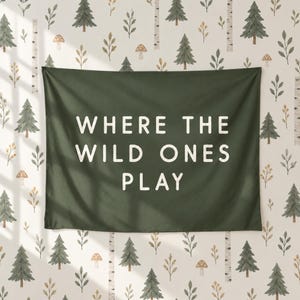 May include: A dark green rectangular wall tapestry featuring the white text "WHERE THE WILD ONES PLAY" in a clean sans-serif font, displayed against a light-coloured background patterned with small green pine trees and brown mushrooms.