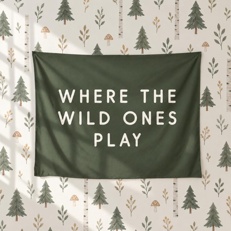 May include: A dark green rectangular wall tapestry featuring the white text "WHERE THE WILD ONES PLAY" in a clean sans-serif font, displayed against a light-coloured background patterned with small green pine trees and brown mushrooms.