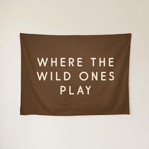 May include: A rectangular brown wall tapestry featuring the text WHERE THE WILD ONES PLAY in a bold, off-white sans-serif font centred on the fabric. The design is simple and clean, with the lettering clearly visible against the dark background.