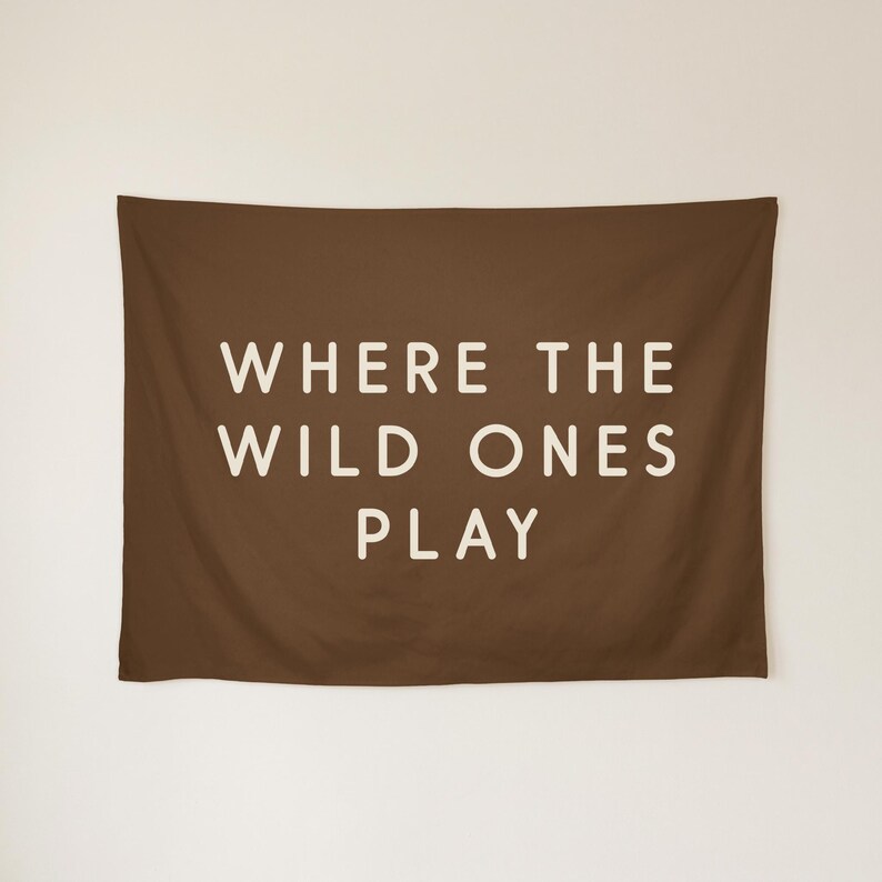 May include: A rectangular brown wall tapestry featuring the text WHERE THE WILD ONES PLAY in a bold, off-white sans-serif font centred on the fabric. The design is simple and clean, with the lettering clearly visible against the dark background.