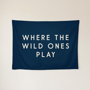May include: A rectangular navy blue wall tapestry featuring the white text WHERE THE WILD ONES PLAY centred in a clean, sans-serif font. The fabric has a smooth, matte finish and is displayed horizontally against a plain background.