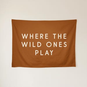 May include: A rectangular fabric wall tapestry in a solid burnt orange colour. Centered on the fabric is the text "WHERE THE WILD ONES PLAY" written in a clean, sans-serif, off-white font across three lines of capitalised lettering.