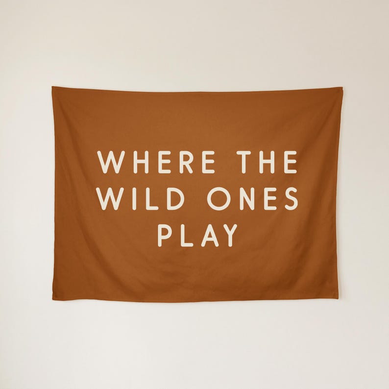 May include: A rectangular fabric wall tapestry in a solid burnt orange colour. Centered on the fabric is the text "WHERE THE WILD ONES PLAY" written in a clean, sans-serif, off-white font across three lines of capitalised lettering.