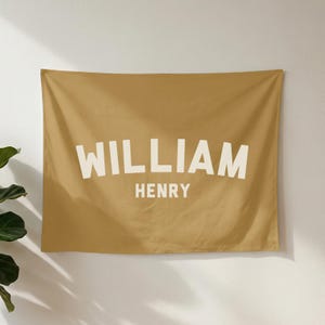 May include: A rectangular mustard yellow fabric wall tapestry featuring the name WILLIAM HENRY printed in large, bold, off-white capital letters. The word WILLIAM is arched across the top, while HENRY is centred below in a straight line.