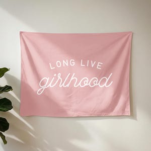 May include: A rectangular wall tapestry in a solid dusty pink colour featuring the white text "LONG LIVE" in a bold sans-serif font above the word "girlhood" written in a white cursive script. The fabric has soft folds and a matte finish.
