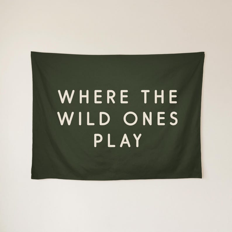 May include: A rectangular dark olive green wall tapestry featuring the white text WHERE THE WILD ONES PLAY centred in a clean sans-serif font. The fabric has a smooth texture and hangs horizontally with a simple, minimalist aesthetic.