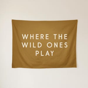 May include: A rectangular mustard yellow wall tapestry featuring the white text WHERE THE WILD ONES PLAY centred in the middle. The fabric has a smooth texture and hangs with soft folds, displaying clear, bold, sans-serif lettering.