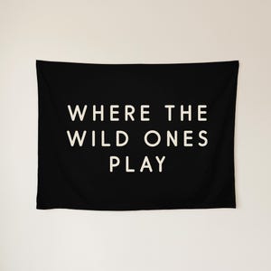 May include: A rectangular black wall tapestry featuring the white text WHERE THE WILD ONES PLAY centred in a clean, sans-serif font. The fabric is smooth and opaque, displaying the bold lettering clearly against the dark background.
