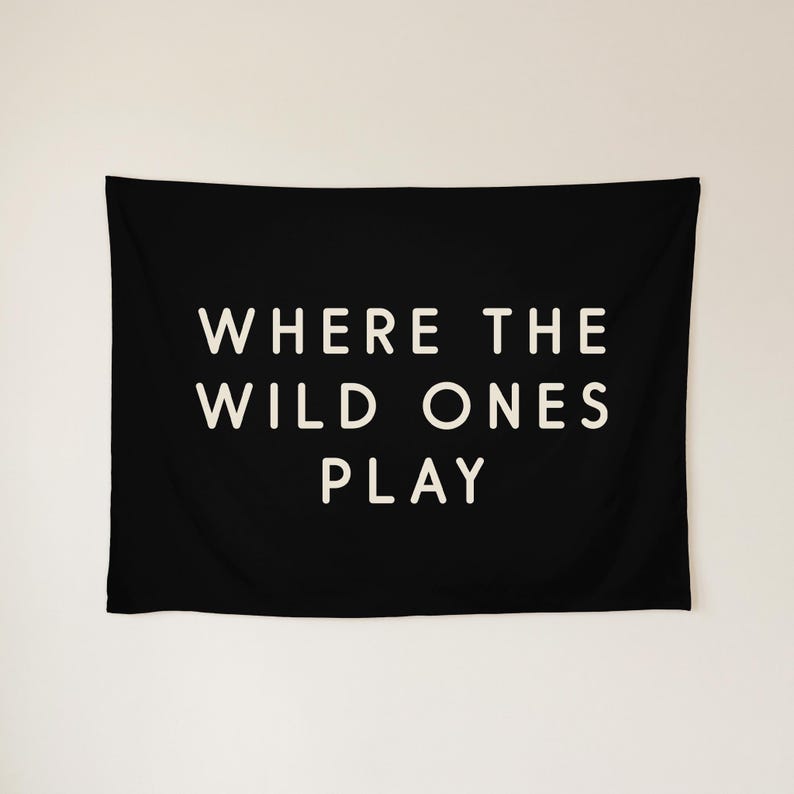 May include: A rectangular black wall tapestry featuring the white text WHERE THE WILD ONES PLAY centred in a clean, sans-serif font. The fabric is smooth and opaque, displaying the bold lettering clearly against the dark background.
