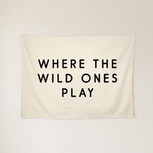 May include: A rectangular off-white wall tapestry featuring the black text WHERE THE WILD ONES PLAY centred in a clean sans-serif font. The fabric has a smooth texture and hangs flat against the wall, displaying the three-line phrase clearly.