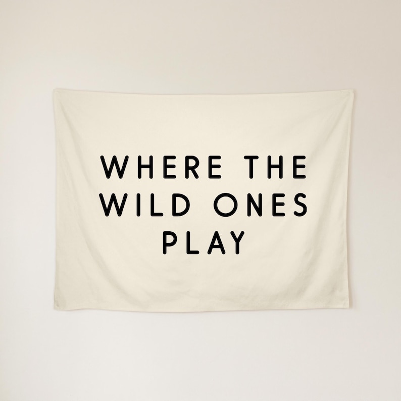 May include: A rectangular off-white wall tapestry featuring the black text WHERE THE WILD ONES PLAY centred in a clean sans-serif font. The fabric has a smooth texture and hangs flat against the wall, displaying the three-line phrase clearly.