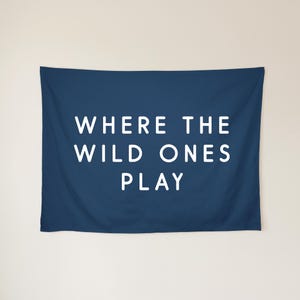 May include: A rectangular navy blue wall tapestry featuring the white text WHERE THE WILD ONES PLAY centred in the middle. The fabric has a smooth finish and is displayed horizontally with clean, straight edges and a solid dark blue background.