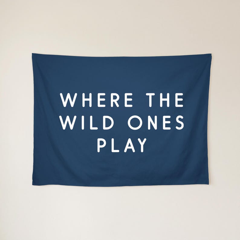 May include: A rectangular navy blue wall tapestry featuring the white text WHERE THE WILD ONES PLAY centred in the middle. The fabric has a smooth finish and is displayed horizontally with clean, straight edges and a solid dark blue background.