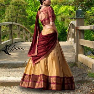 May include: A traditional Indian outfit featuring a gold-colored skirt with a burgundy border and a matching burgundy shawl. The top is a burgundy color with gold accents. The outfit is worn on a wooden bridge with a green background.