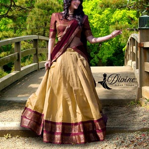May include: A traditional Indian outfit featuring a gold skirt with a burgundy border, paired with a burgundy top and matching shawl. The outfit is worn outdoors on a wooden bridge, with the text "Divine Gleam Trends".