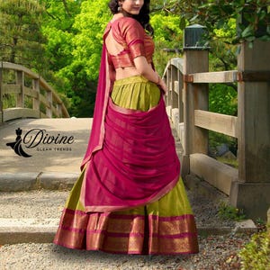 May include: A traditional Indian outfit featuring a green skirt with a gold and magenta border, a magenta draped shawl, and a red-orange blouse. The outfit is worn outdoors near a bridge with the text "Divine Gleam Trends".