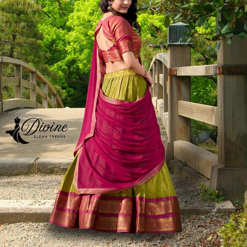 May include: A traditional Indian outfit featuring a green skirt with a gold and magenta border, a magenta draped shawl, and a red-orange blouse. The outfit is worn outdoors near a bridge with the text "Divine Gleam Trends".