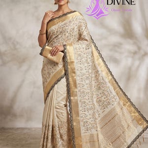 May include: Cream-colored sari with gold borders and black lace trim. The sari features a repeating pattern of small figures and shapes. A matching clutch is held in the model's hand. The Divine Gloom Trends logo is visible in the upper right corner.