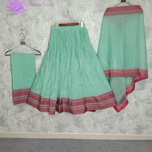 May include: A three-piece mint green outfit, including a skirt and a shawl, hangs on a wall. The skirt and shawl have a decorative border with red and brown accents. The outfit is from the brand "DIVINE Global Trends".