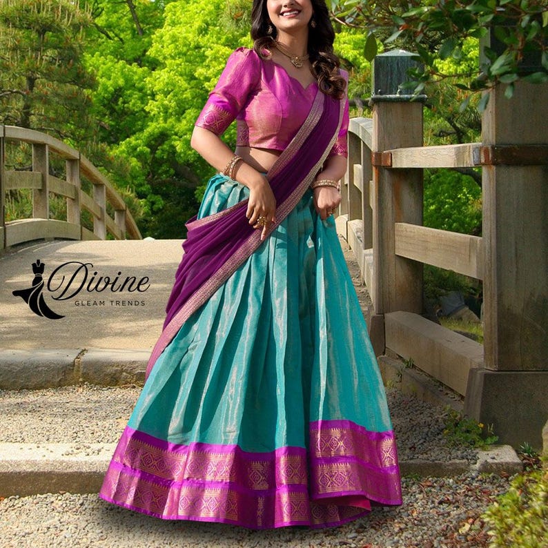 May include: A traditional Indian outfit featuring a teal skirt with a magenta and gold border, paired with a magenta blouse and a purple shawl. The outfit is embellished with gold accents and intricate patterns. The brand logo "Divine Gleam Trends" is visible.