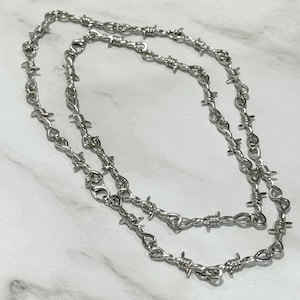 以下が含まれることがあります： 有刺鉄線デザインのシルバーカラーのネックレス。ネックレスは金属製で、チェーンに沿って一連の小さくて鋭いバリが付いています。ネックレスは白とグレーの大理石の背景に対して表示されています。