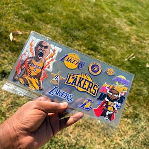 May include: A clear plastic sheet filled with various Los Angeles Lakers stickers. The stickers feature the Lakers logo, basketballs, and cartoon illustrations of players in yellow and purple uniforms. The background is green grass.