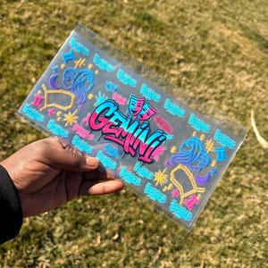 Puede incluir: Una cartera de plástico transparente con un diseño colorido de Géminis. El diseño presenta la palabra "Gemini" en estilo graffiti, junto con símbolos del zodiaco y estrellas en rosa, azul y amarillo.