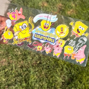 Può includere: Un foglio rettangolare trasparente con diversi adesivi colorati dei personaggi SpongeBob SquarePants e Patrick Stella. Il design centrale include il testo SpongeBob SquarePants in blu e giallo, con una piccola graffetta attaccata in alto.